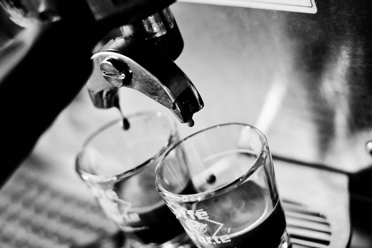 Black and white close-up of espresso dripping from a metal portafilter into two small clear espresso glasses on a drip tray.